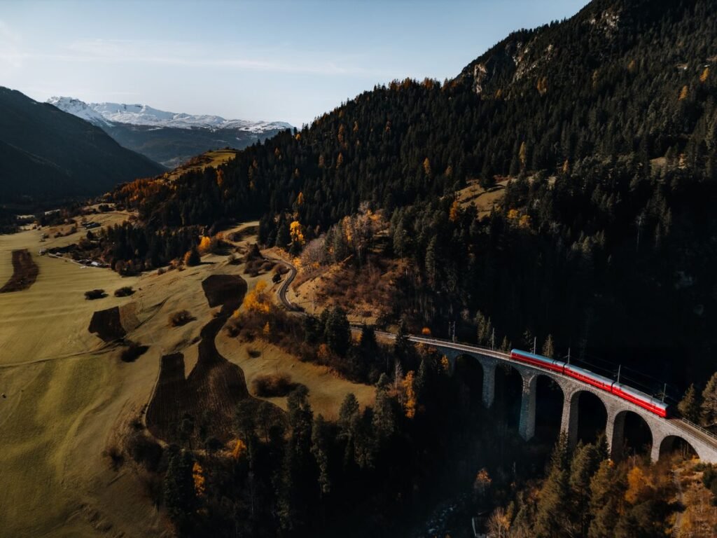Red train crossing a curved stone viaduct in an autumn mountain valley