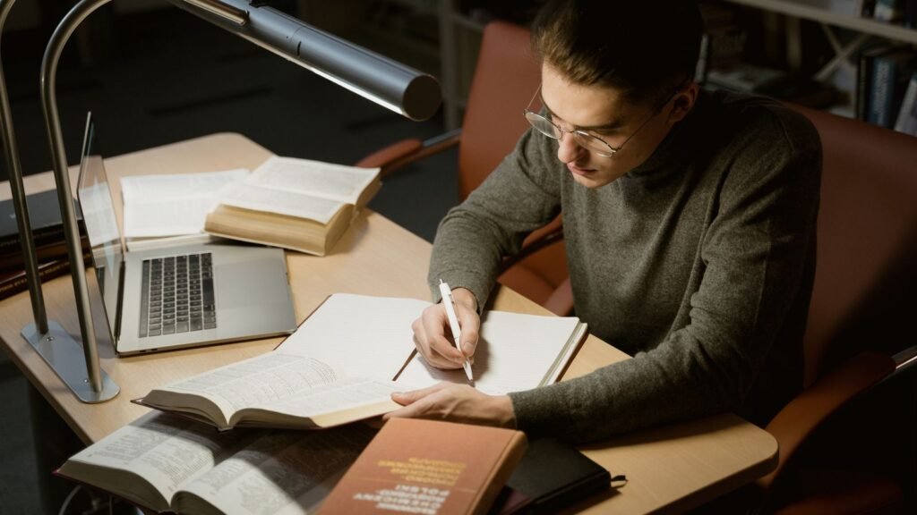 Student taking notes beside an open laptop and stacked reference books at a library desk