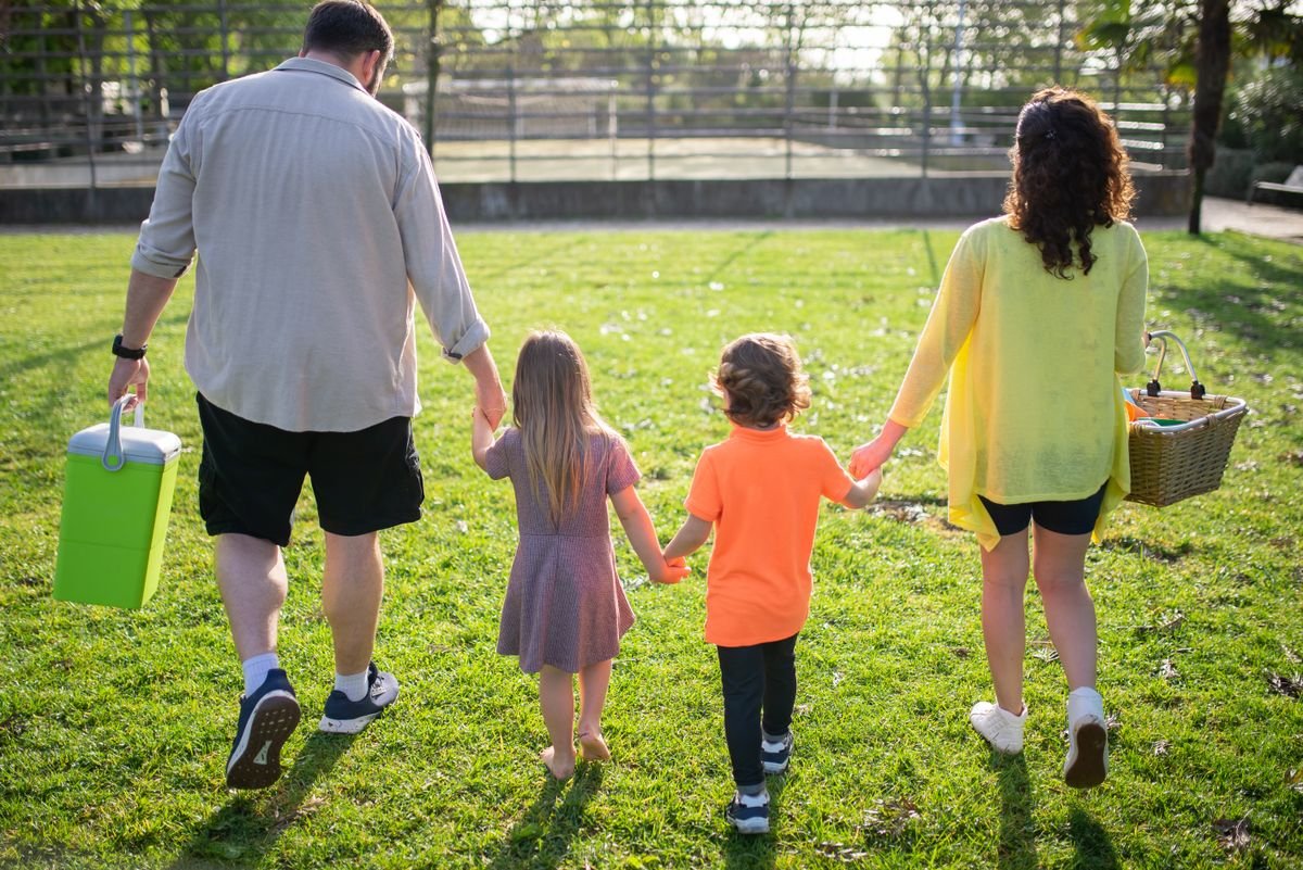 Family walking across a park carrying a green cooler and picnic basket