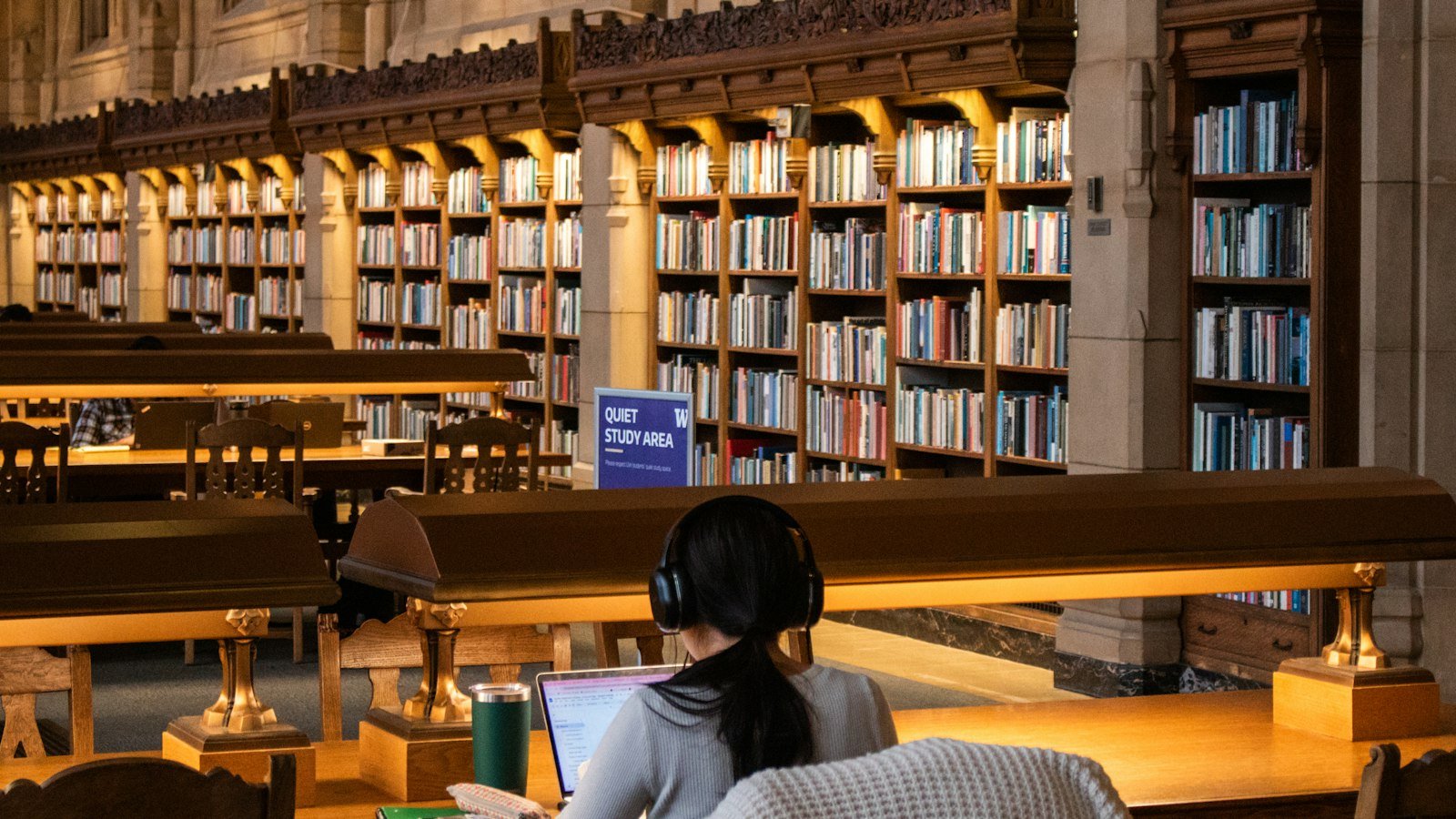 Student at a laptop in a university library reading room lined with bookshelves